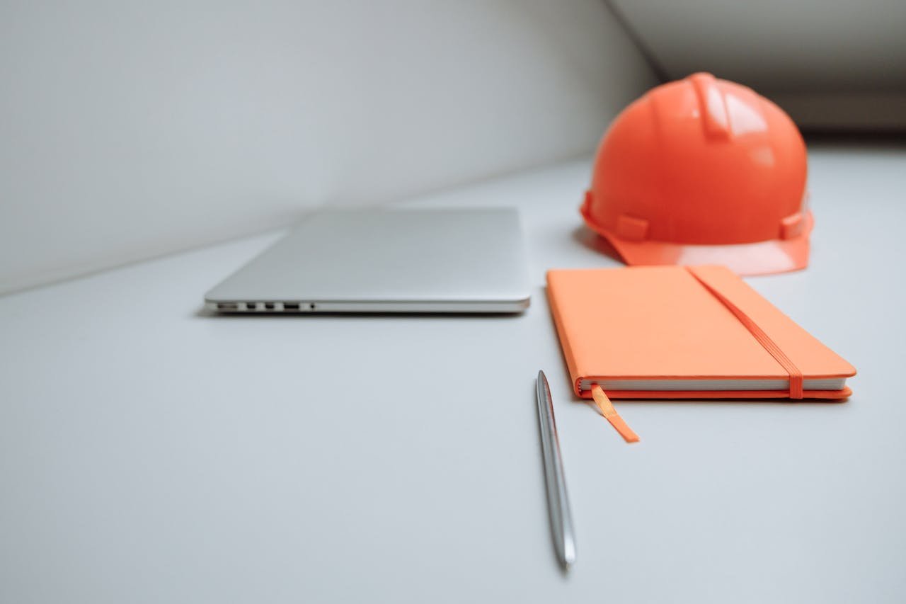 A minimalist office setup featuring a laptop, orange notebook, and safety hard hat.